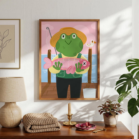 May include: A framed illustration depicts a green frog wearing a straw hat and overalls, holding a pink fish with an X mark. The frog stands on a wooden dock with a fishing rod, a seagull, and a bucket. The background features a pink sky and blue water.