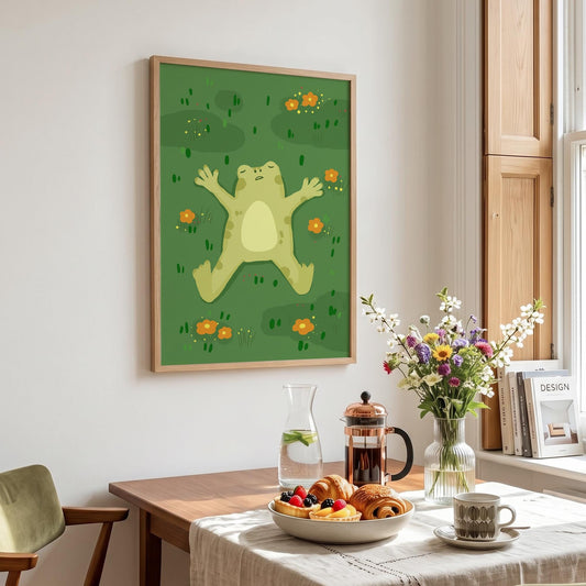 May include: A framed art print featuring a cartoon frog lying on its back in a field of green grass and orange flowers. The frog is light green with a yellow belly. The frame is a light wood color.