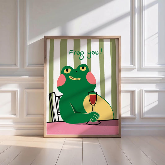 May include: A framed art print featuring a cartoon frog holding a glass of red wine. The frog is green with pink cheeks and yellow eyes, seated at a table with the text "Frog you!" above it. The background has green and white stripes.