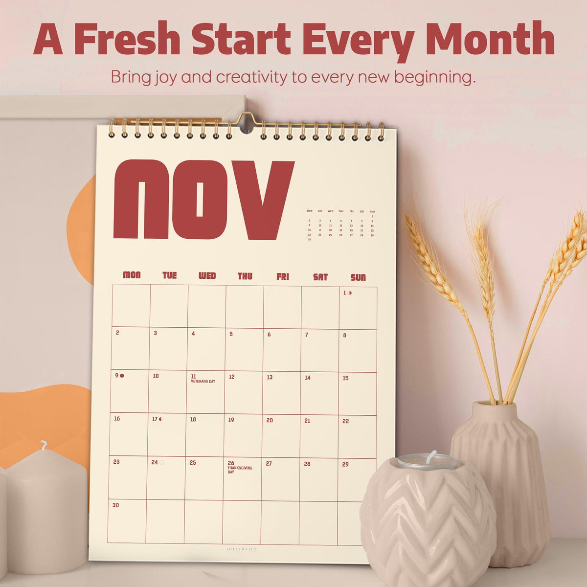 May include: A wall calendar for November with a gold spiral binding. The calendar has a cream-colored background with red text that reads "NOV". The top of the calendar reads "A Fresh Start Every Month". A vase with wheat stalks and a candle holder are in the foreground.