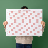 May include: A white wall calendar titled 26/27 YEAR PLANNER with months listed from October to September. The grid features alternating white and dusty pink blocks representing days of the week, with the brand name JOLIEDAILY at the bottom.