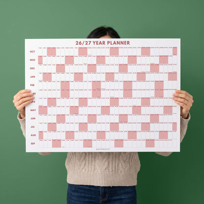 May include: A white wall calendar titled 26/27 YEAR PLANNER with months listed from October to September. The grid features alternating white and dusty pink blocks representing days of the week, with the brand name JOLIEDAILY at the bottom.
