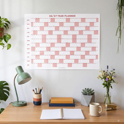 May include: A wall-mounted 26/27 year planner featuring a grid layout with alternating white and dusty pink blocks. The calendar displays months from October to September, with the text 26/27 YEAR PLANNER printed in red at the top center.