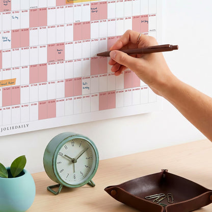 May include: A wall-mounted annual calendar with pink and white grid sections, featuring the text JOLIEDAILY. A round, light green analog clock with a white face and black numerals sits next to a dark brown leather valet tray containing paper clips.