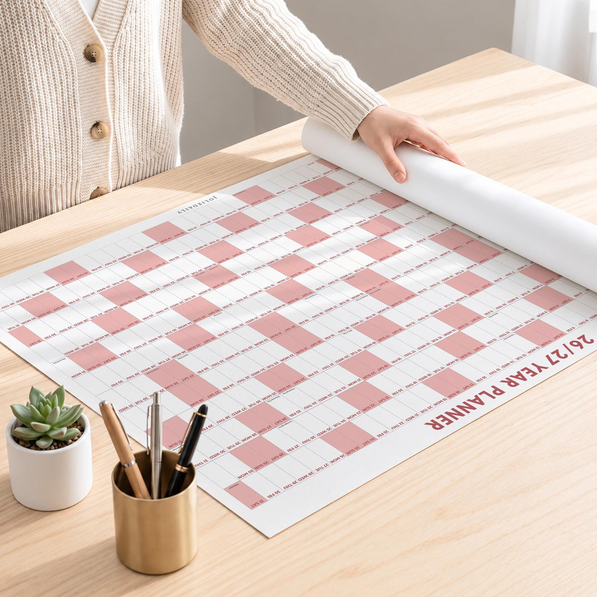 May include: A large wall calendar titled 26/27 YEAR PLANNER with a white and dusty pink checkered grid layout. The planner is partially rolled up on one side. A small white ceramic pot with a green succulent and a gold pen holder with three pens.