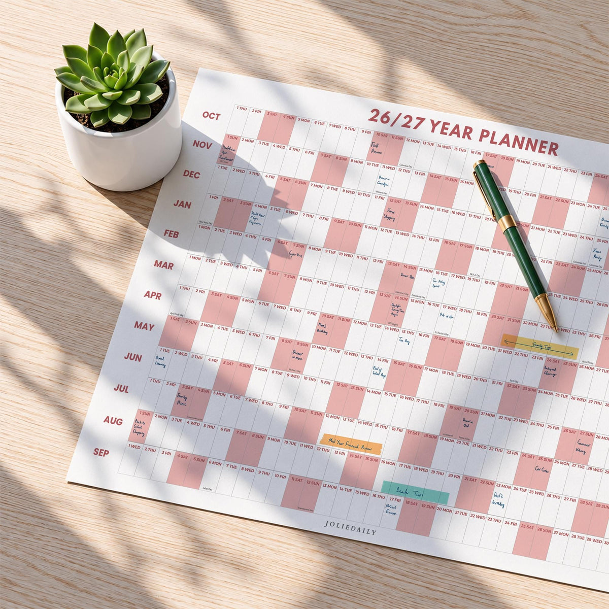 May include: A 26/27 year planner wall calendar featuring a grid layout with alternating white and dusty pink blocks, months listed from October to September, a green and gold pen resting on the surface, and a small succulent in a white pot.