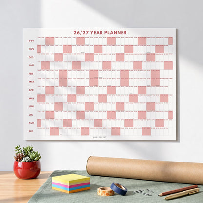 May include: A wall-mounted 26/27 year planner featuring a grid layout with alternating white and dusty pink columns. The months from October to September are listed vertically on the left side, with the text 26/27 YEAR PLANNER at the top.