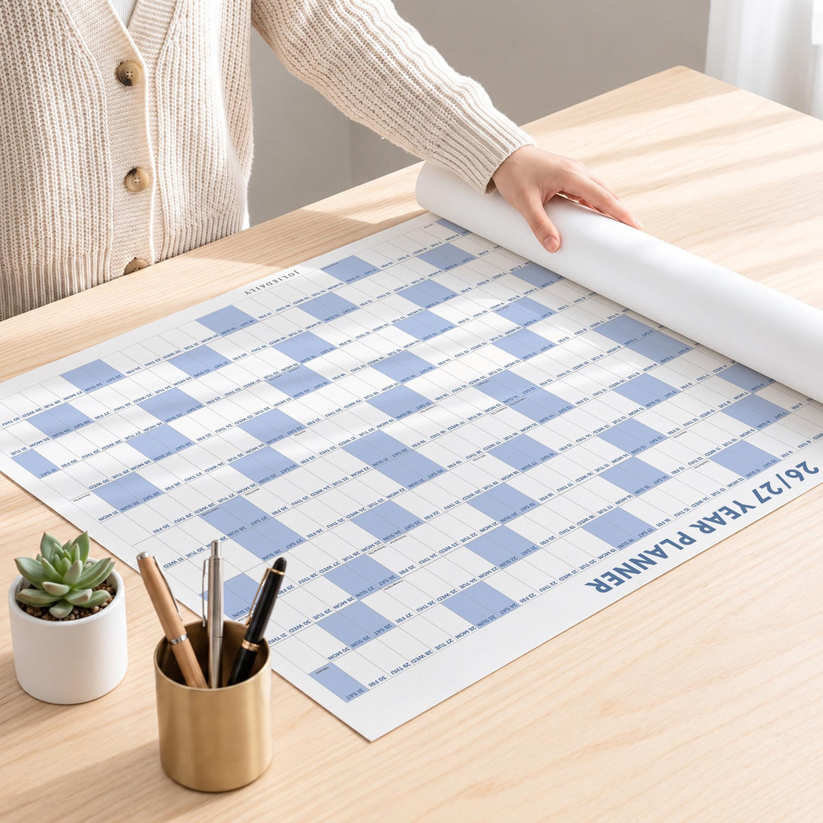 May include: A large wall calendar titled 26/27 YEAR PLANNER with a blue and white checkered grid pattern. The planner is partially rolled up on a wooden surface next to a small potted succulent and a metallic pen holder containing three pens.