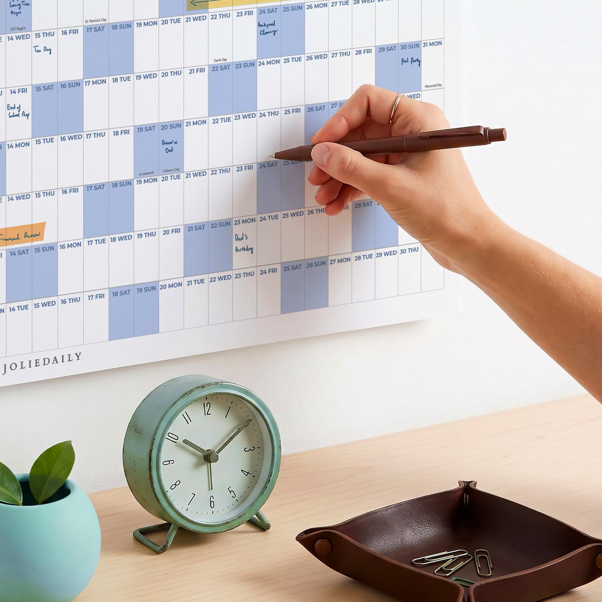 May include: A wall calendar with a grid layout, light blue and white blocks, and the text JOLIEDAILY. A round, mint green analog clock with a stand, a dark brown leather tray containing metal paper clips, and a small teal planter are present.