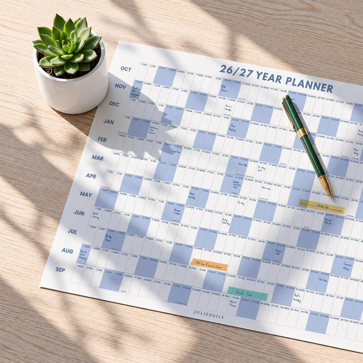 May include: A 26/27 year planner wall calendar with a grid layout featuring alternating white and light blue blocks for months and days. The planner includes handwritten notes, a green and gold pen resting on the surface, and a small white pot with a green succulent.