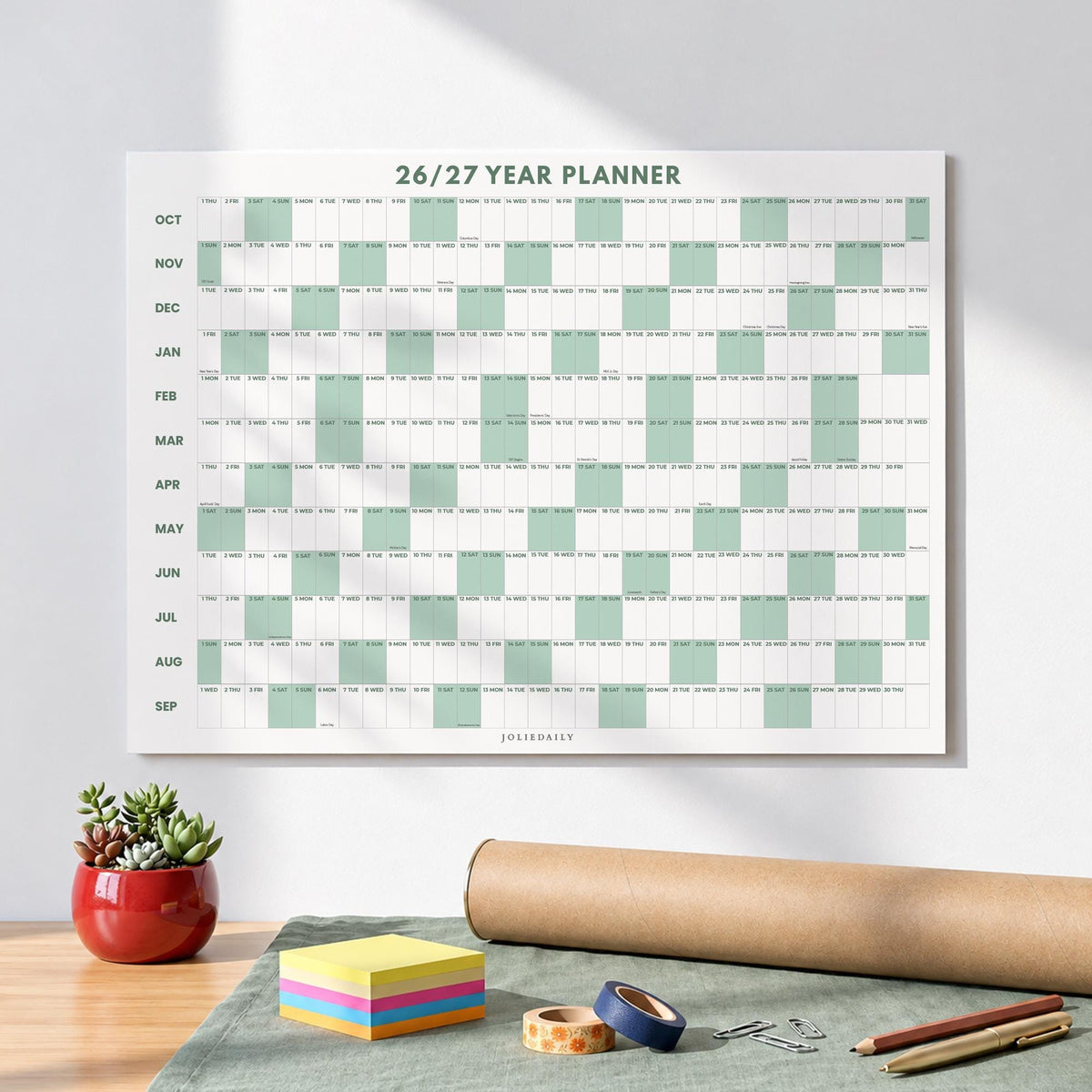 May include: A wall-mounted 26/27 year planner calendar featuring a grid layout with alternating white and sage green columns. The months from October to September are listed vertically on the left side, with the text JOLIEDAILY at the bottom.