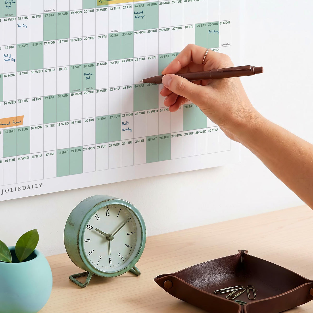 May include: A wall-mounted annual calendar with sage green and white grid sections, featuring the text JOLIEDAILY. A round, sage green analog clock with a white face and black hands sits next to a dark brown leather tray containing metal paperclips.