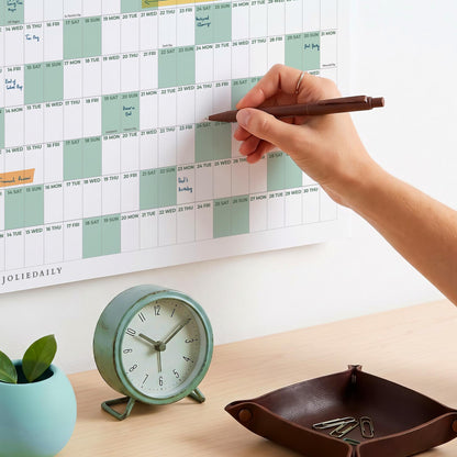 May include: A wall-mounted annual calendar with sage green and white grid sections, featuring the text JOLIEDAILY. A round, sage green analog clock with a white face and black hands sits next to a dark brown leather tray containing metal paperclips.