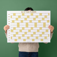 May include: A white wall calendar titled 26/27 YEAR PLANNER with gold text. The grid features rows for months from October to September, with alternating white and gold columns representing days. The bottom center includes the brand name JOLIEDAILY.