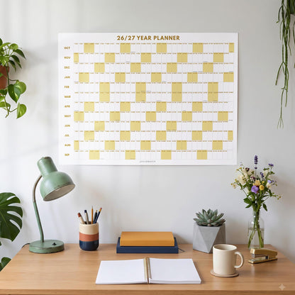 May include: A wall-mounted 26/27 year planner featuring a grid layout with alternating white and mustard yellow blocks. The calendar displays months from October to September with clear text headings and a clean, organized design for tracking dates.