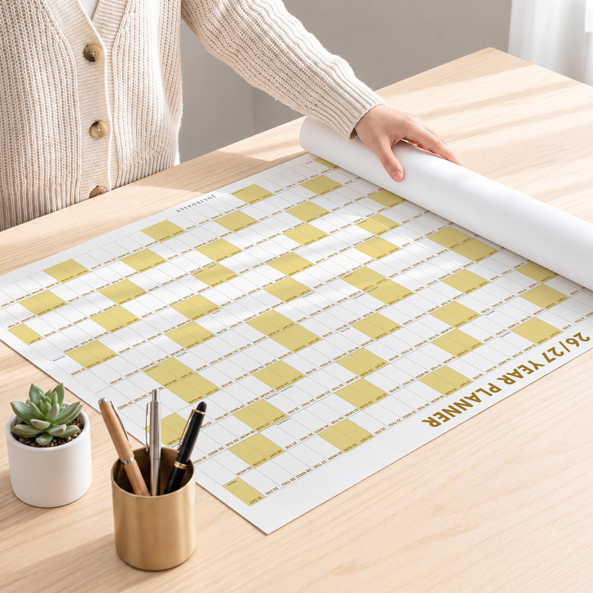 May include: A large white wall calendar titled 26/27 YEAR PLANNER with a grid of yellow and white squares. The planner is partially rolled, featuring small text for dates and days of the week. A small succulent in a white pot and a gold pen holder are present.