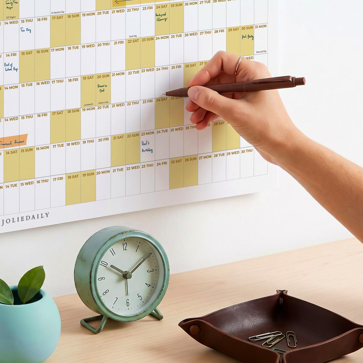 May include: A wall calendar with a grid layout featuring alternating white and pale yellow columns, marked with the text JOLIEDAILY. A brown pen is held against the calendar, while a round mint green analog clock and a brown leather tray sit below.