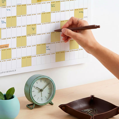 May include: A wall calendar with a grid layout featuring alternating white and pale yellow columns, marked with the text JOLIEDAILY. A brown pen is held against the calendar, while a round mint green analog clock and a brown leather tray sit below.