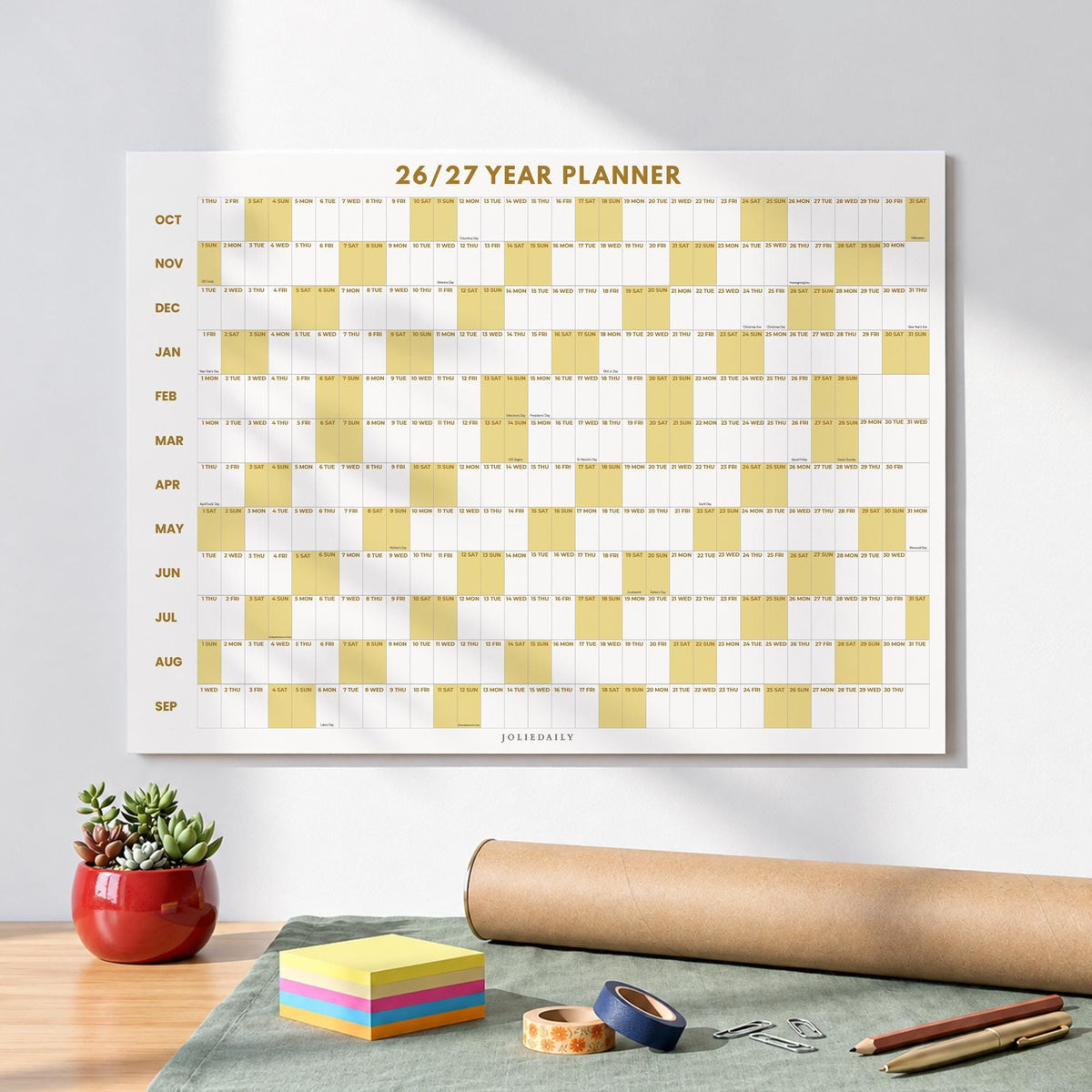 May include: A wall-mounted 26/27 year planner with a white background, gold text, and alternating white and yellow grid columns. The months from October to September are listed vertically on the left side, with the brand name JOLIEDAILY at the bottom.