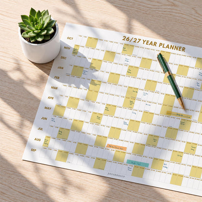 May include: A 26/27 year planner wall calendar with a grid layout featuring alternating white and yellow blocks for months and days. The planner includes a green pen resting on the surface and a small green succulent in a white ceramic pot.