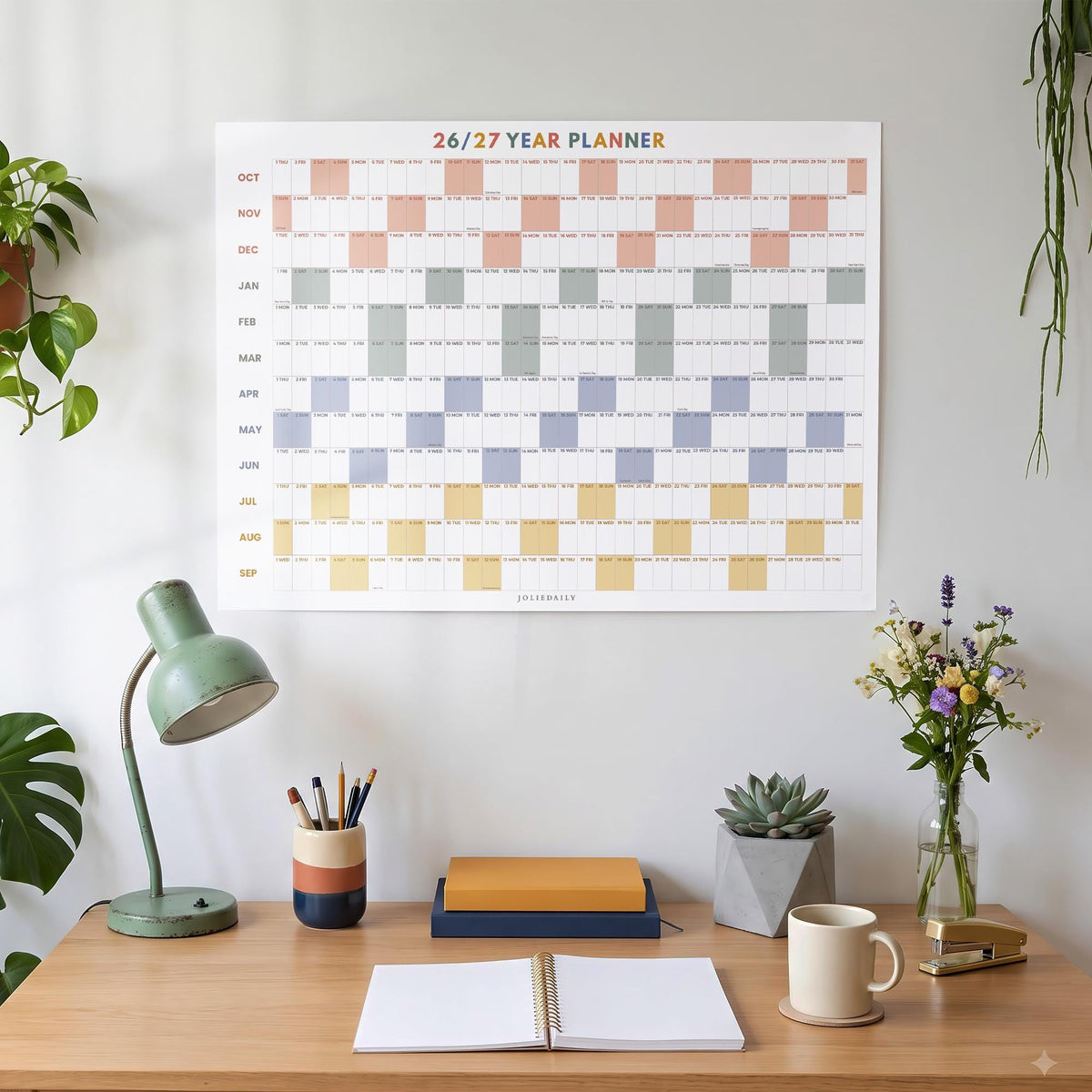 May include: A wall-mounted 26/27 year planner featuring a grid layout with horizontal rows of months in alternating pastel colors including peach, sage green, periwinkle blue, and mustard yellow, with the text 26/27 YEAR PLANNER at the top.