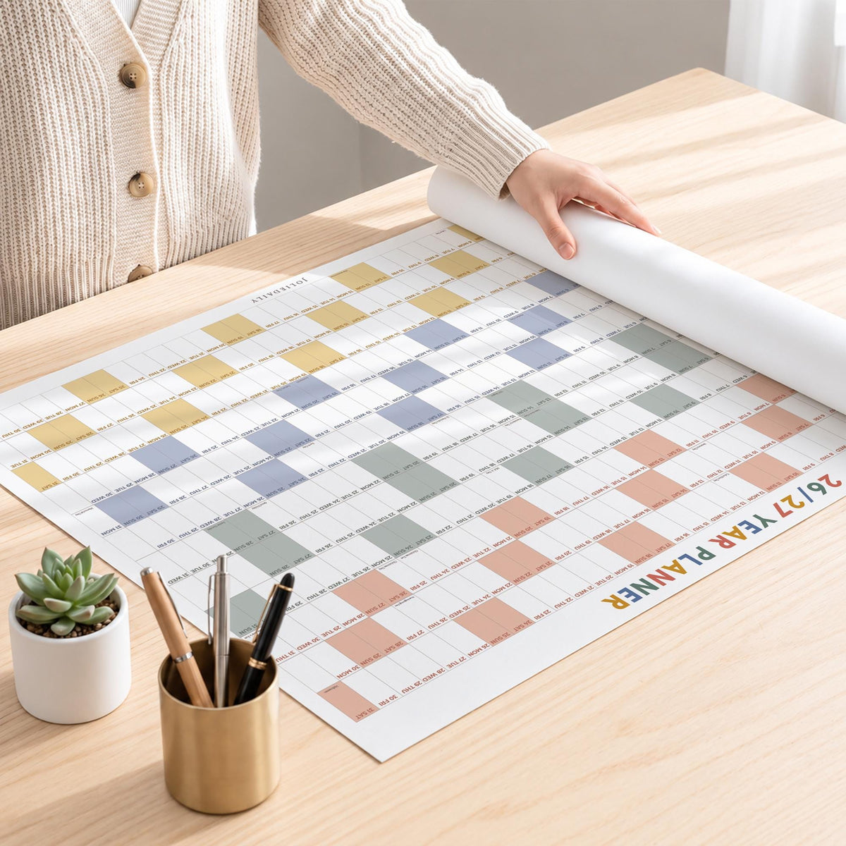 May include: A large wall calendar titled 26/27 YEAR PLANNER with a grid of colored squares in yellow, blue, green, and peach. The planner is partially rolled on a wooden surface next to a gold pen holder containing three pens and a small potted succulent.