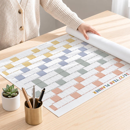 May include: A large wall calendar titled 26/27 YEAR PLANNER with a grid of colored squares in yellow, blue, green, and peach. The planner is partially rolled on a wooden surface next to a gold pen holder containing three pens and a small potted succulent.