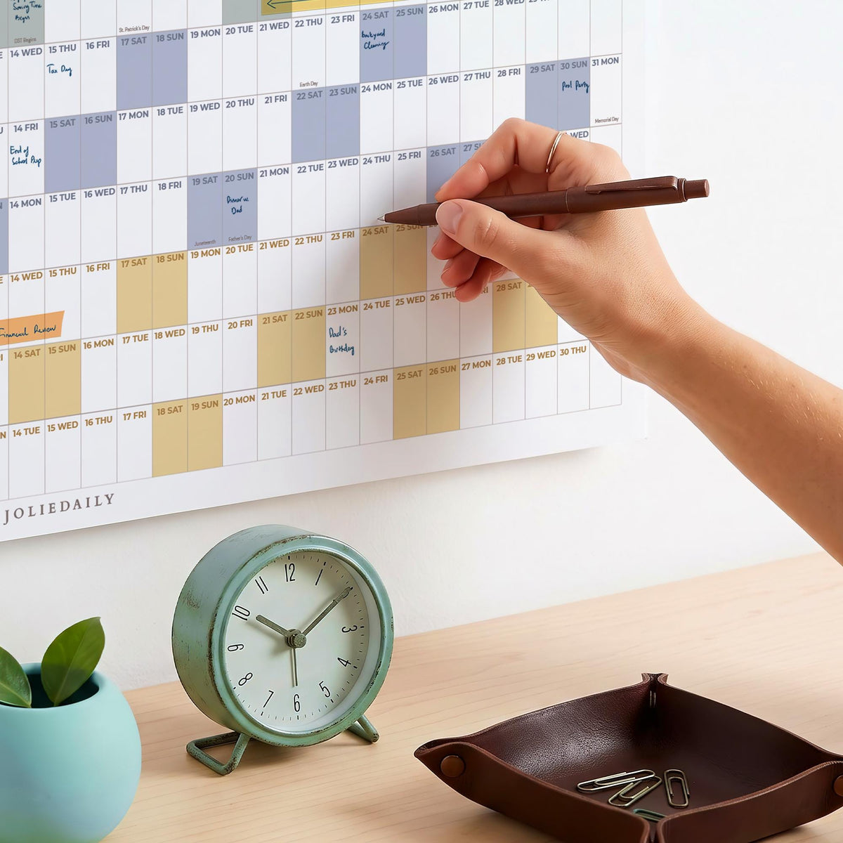 May include: A wall calendar with a grid layout featuring blue and yellow highlighted dates and the text JOLIEDAILY, a round mint green analog clock with a white face, and a dark brown leather tray containing several silver metal paper clips.