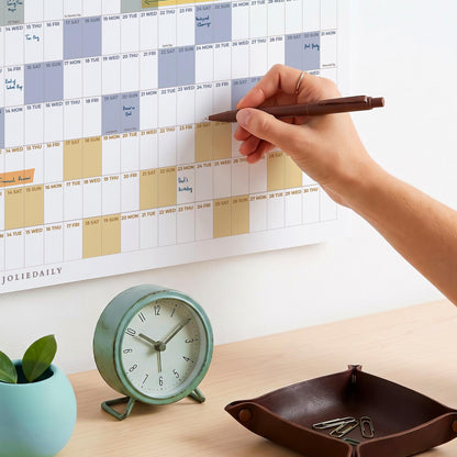 May include: A wall calendar with a grid layout featuring blue and yellow highlighted dates and the text JOLIEDAILY, a round mint green analog clock with a white face, and a dark brown leather tray containing several silver metal paper clips.
