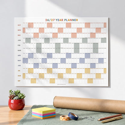 May include: A wall-mounted 26/27 year planner calendar featuring a grid layout with months labeled from October to September. The design includes alternating colored blocks in peach, gray, blue, and yellow, with the text 26/27 YEAR PLANNER.