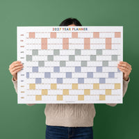 May include: A 2027 year planner wall calendar featuring a grid layout with months listed vertically from January to December. The design uses alternating colored blocks in shades of peach, gray, blue, and yellow to distinguish days and weeks.