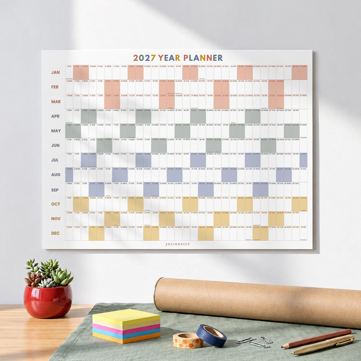 May include: A 2027 year planner wall calendar featuring a grid layout with months listed vertically from January to December. The design uses alternating blocks of peach, gray, blue, and yellow colors to organize the days of the year.