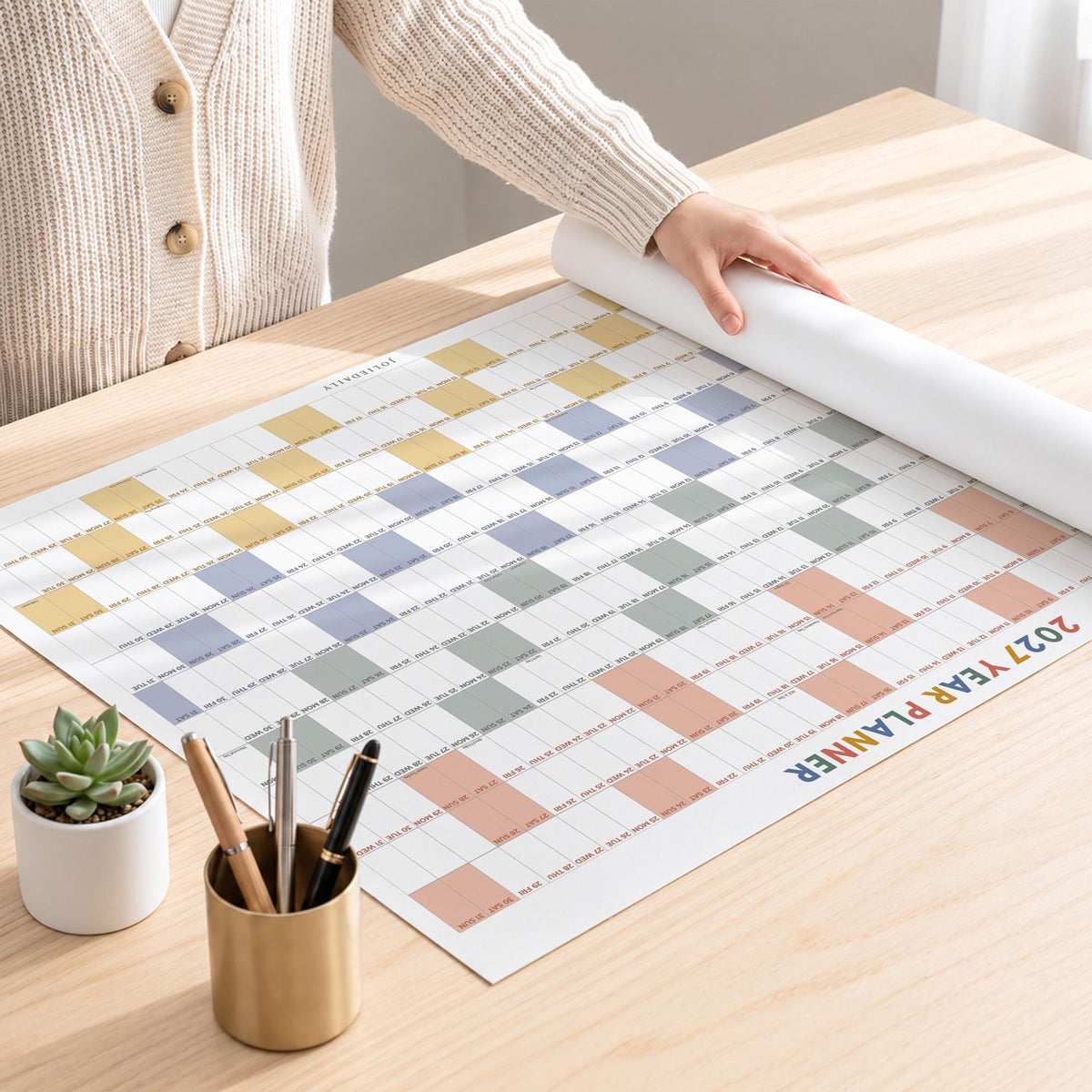 May include: A 2027 year planner wall calendar featuring a grid layout with alternating rows of yellow, blue, green, and peach colored squares. The planner is printed on white paper and includes a small potted succulent and a gold pen holder.