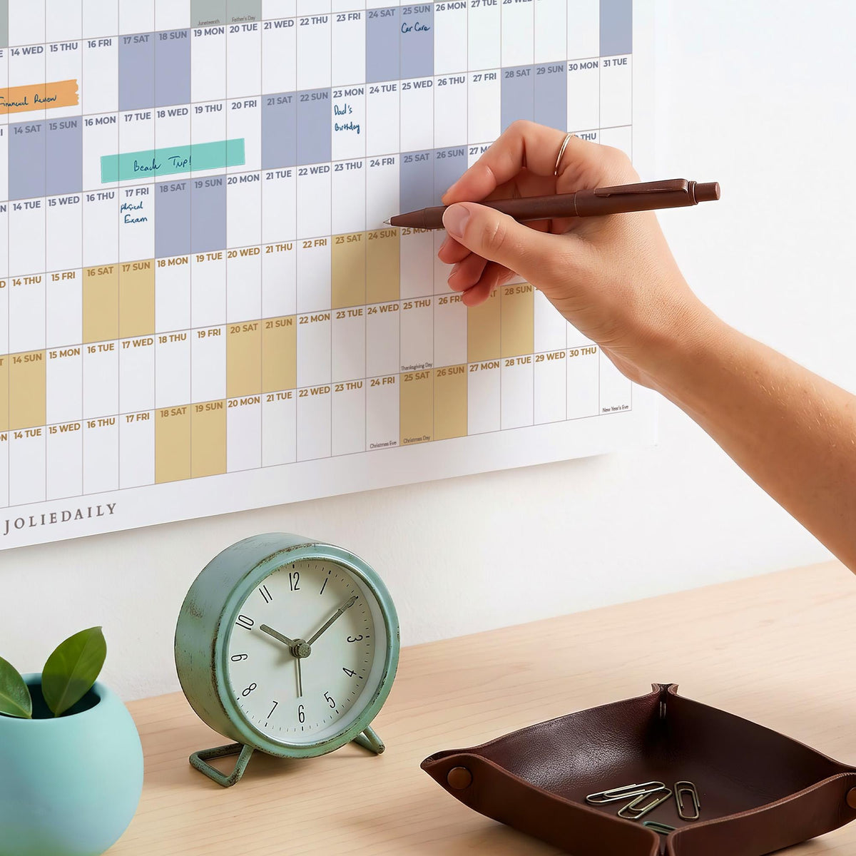 May include: A wall calendar with a grid layout featuring blue, yellow, and orange color-blocked sections, labeled JOLIEDAILY. A brown pen is held over the calendar. A mint green analog clock and a dark brown leather tray with paperclips are present.