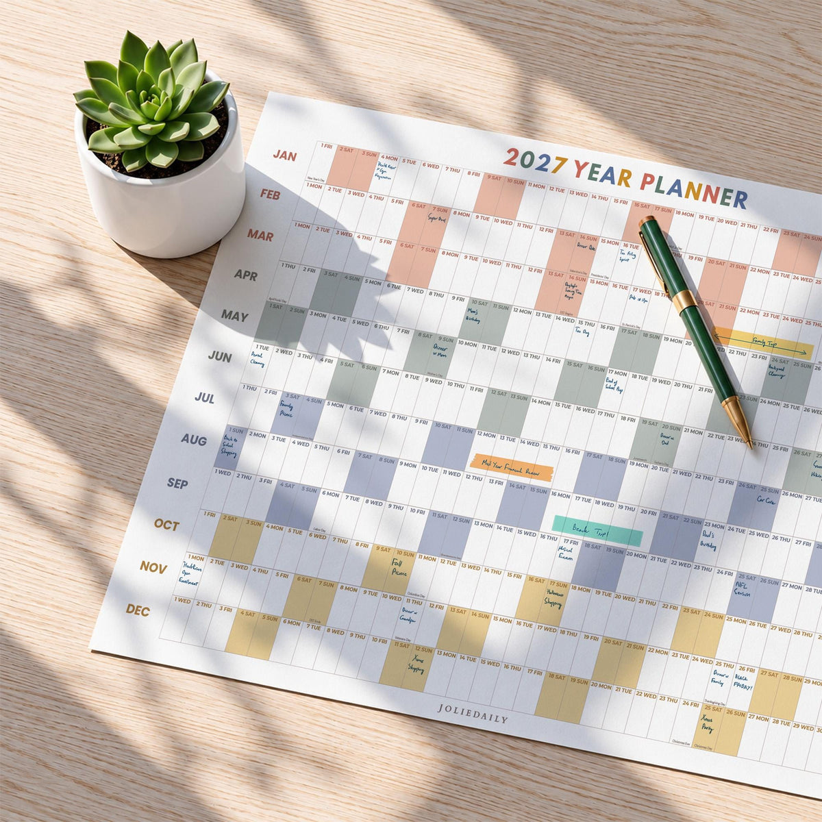 May include: A 2027 year planner wall calendar with a grid layout featuring months from January to December in rows. The planner has color-coded blocks for days and includes the text 2027 YEAR PLANNER and JOLIEDAILY. A green pen rests on the paper.