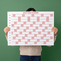 May include: A 2027 year planner wall calendar featuring a horizontal grid layout. The design includes white and soft pink rectangular blocks representing days of the month, with months listed vertically on the left and the text JULIEDAILY at the bottom.