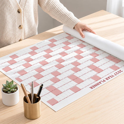 May include: A 2027 year planner wall calendar featuring a white and dusty pink checkered grid pattern. The planner is laid out flat on a surface, with a metallic pen holder containing three pens and a small potted succulent plant nearby.