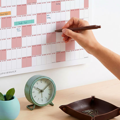 May include: A white wall calendar with pink shaded columns and the text JOLIEDAILY, a round mint green analog clock with a stand, a dark brown leather valet tray containing metal paper clips, and a dark brown pen writing on the calendar.