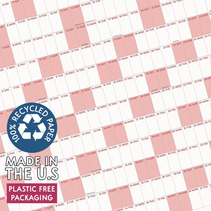 May include: A wall calendar featuring a grid layout with alternating white and light pink columns. The calendar includes printed dates and days of the week. A blue circular badge displays the text 100% RECYCLED PAPER, MADE IN THE U.S, PLASTIC FREE PACKAGING.