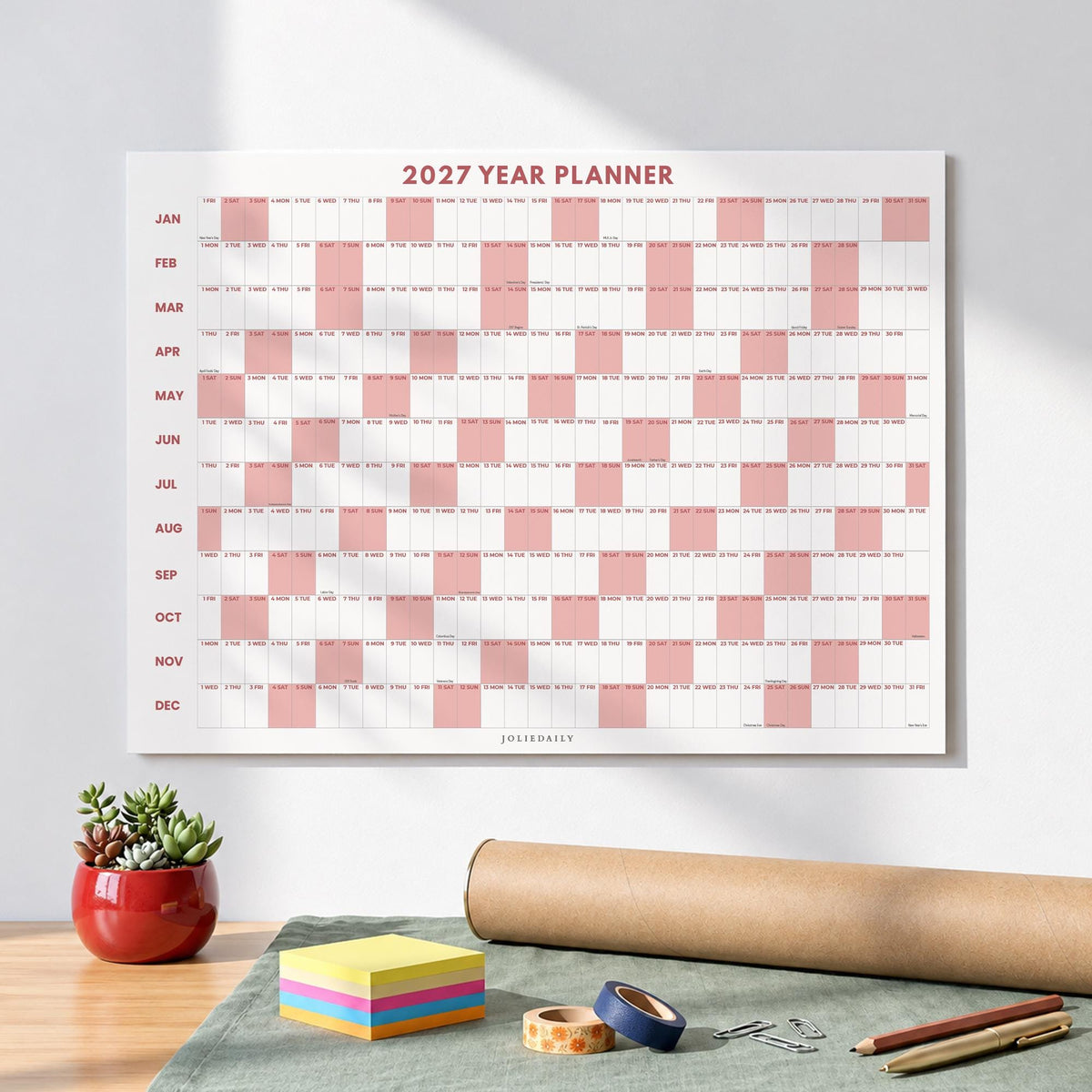 May include: A 2027 year planner wall calendar featuring a grid layout with alternating white and dusty rose pink blocks for each day of the year. The months from January to December are listed vertically on the left side in dark red text.