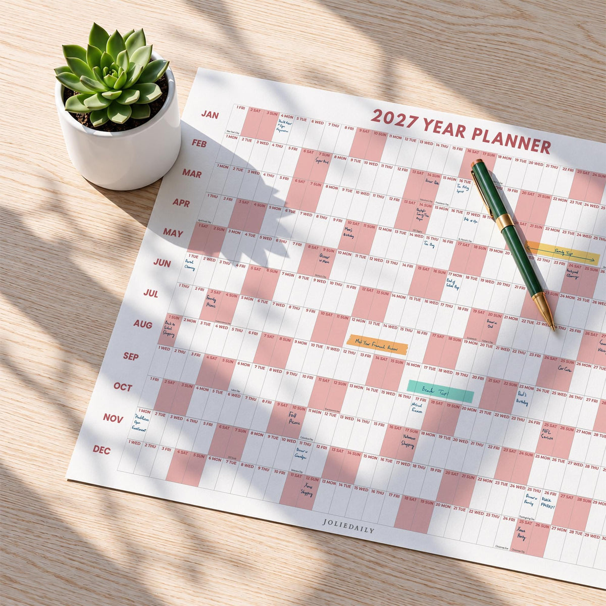 May include: A 2027 year planner wall calendar featuring a grid layout with alternating white and dusty pink blocks for each month. A green and gold pen rests on the paper, and a small green succulent in a white ceramic pot sits nearby.