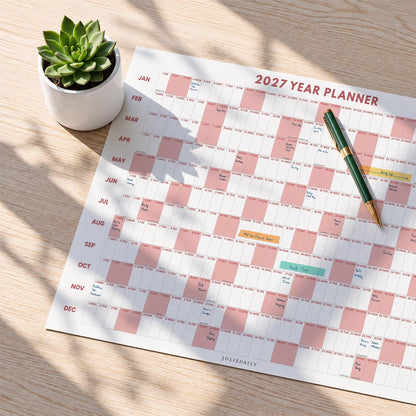 May include: A 2027 year planner wall calendar featuring a grid layout with alternating white and dusty pink blocks for each month. A green and gold pen rests on the paper, and a small green succulent in a white ceramic pot sits nearby.