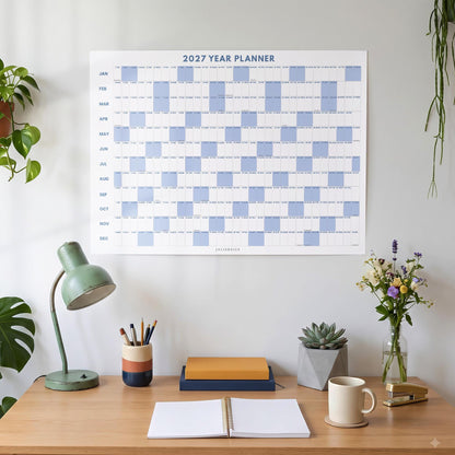May include: A 2027 year planner wall calendar featuring a grid layout with alternating light blue and white blocks for each month from January to December. The text 2027 YEAR PLANNER is printed in blue at the top center of the white paper sheet.