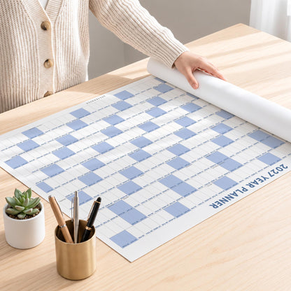 May include: A 2027 year planner wall calendar featuring a grid layout with alternating white and light blue squares. The planner is partially rolled at the top edge and displays the text 2027 YEAR PLANNER in dark blue capital letters.
