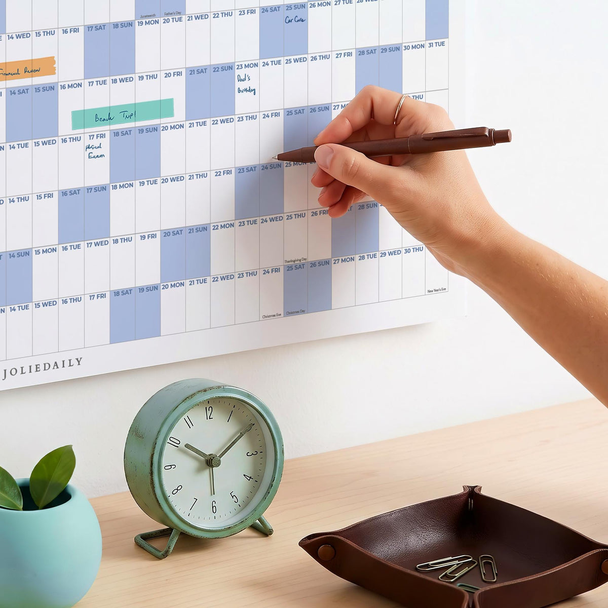 May include: A wall calendar with a grid layout, light blue and white sections, and the text JOLIEDAILY. A round, light green analog clock with a white face and black numbers sits below, alongside a dark brown leather tray containing metal paper clips.