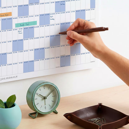 May include: A wall calendar with a grid layout, light blue and white sections, and the text JOLIEDAILY. A round, light green analog clock with a white face and black numbers sits below, alongside a dark brown leather tray containing metal paper clips.