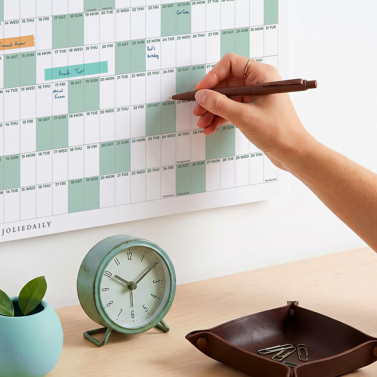 May include: A wall-mounted annual calendar with white and sage green grid sections, featuring handwritten notes. A round sage green analog clock, a dark brown leather valet tray with metal paperclips, and a dark brown pen are also present.