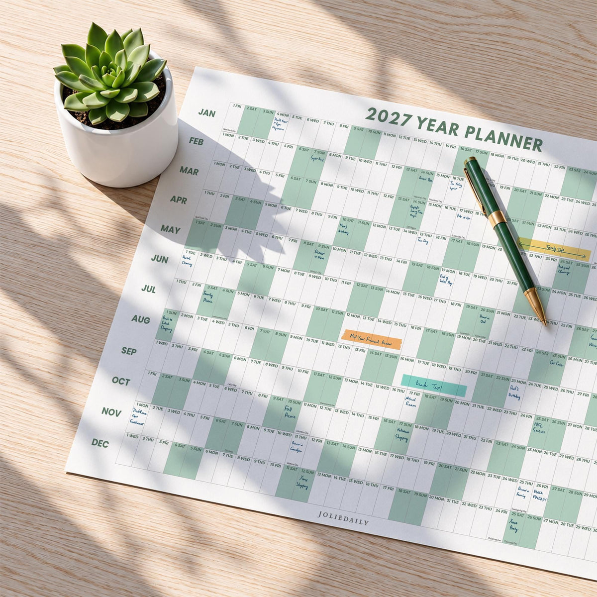 May include: A 2027 year planner wall calendar with a grid layout featuring alternating white and light green columns for each month. The planner includes the text 2027 YEAR PLANNER and JOLIEDAILY. A green and gold pen rests on the paper surface.