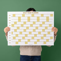 May include: A 2027 year planner wall calendar featuring a white background with gold-colored grid blocks for each month from January to December. The text 2027 YEAR PLANNER is printed in gold at the top, and the brand name JULIEDAILY is at the bottom.