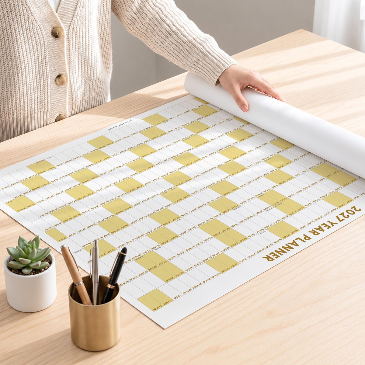 May include: A 2027 year planner wall calendar featuring a grid layout with alternating white and mustard yellow blocks. The planner is printed on a large sheet of paper, partially rolled at the top, and includes a small succulent in a white pot.